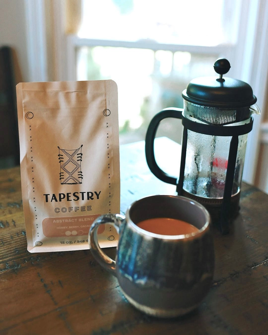 12 oz bag of abstract blend on wood table with french press and mug of coffee