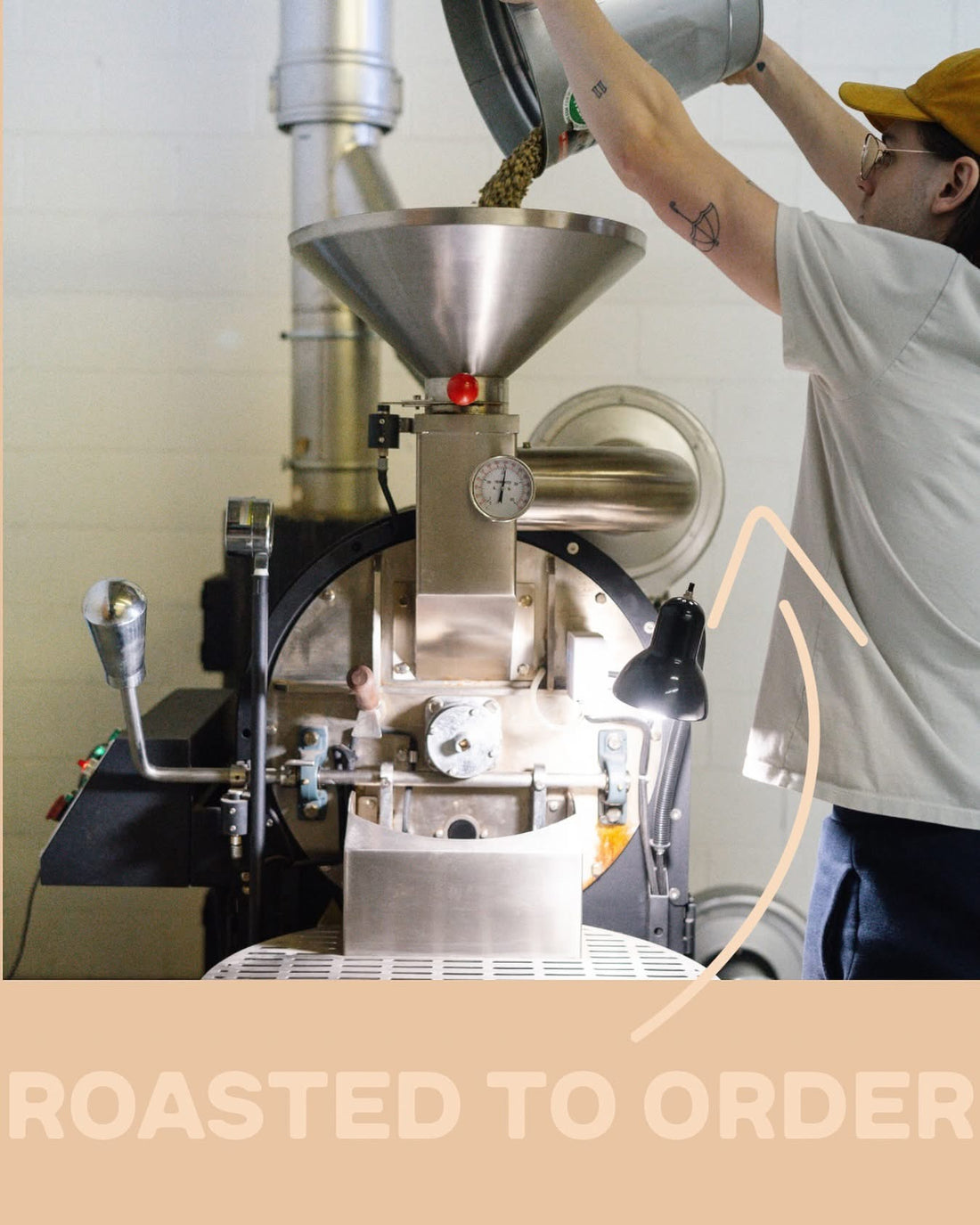 A person in a white t-shirt and yellow cap pours coffee beans into a large stainless steel coffee roaster in a workshop setting. A beige banner at the bottom reads 'ROASTED TO ORDER'.