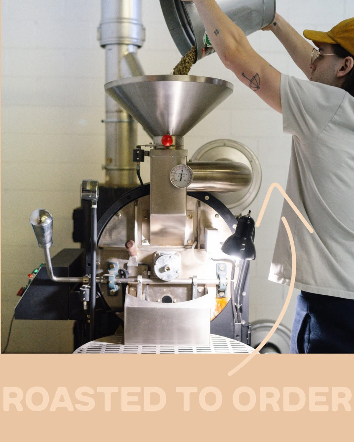 A person in a white t-shirt and yellow cap pours coffee beans into a large stainless steel coffee roaster in a workshop setting. A beige banner at the bottom reads 'ROASTED TO ORDER'.