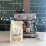 A bag of Tapestry Coffee Abstract Blend stands in front of a stainless steel Frizzino espresso machine on a marble countertop, with a green tiled kitchen wall in the background.