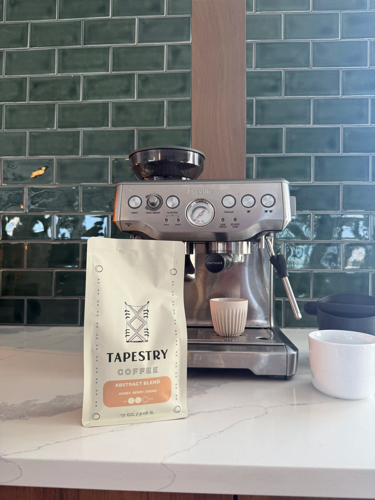 A bag of Tapestry Coffee Arabica Espresso Roast standing on a white marble countertop in front of a stainless steel espresso machine, with a green subway tile backsplash behind it.