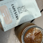 12 oz bag of tapestry coffee abstract blend on wood table with glass of iced coffee
